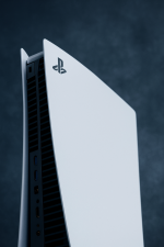 Das Bild zeigt eine PlayStation 5 in einem eleganten, modernen Design. Die Konsole ist überwiegend in Weiß gehalten, mit einer markanten, futuristischen Form, die sich nach oben hin verjüngt. An der Vorderseite sind verschiedene Anschlüsse sichtbar, während die Kanten durch eine glatte Oberfläche und eine leicht abgerundete Form betont werden. Die charakteristischen Lüftungsschlitze an der Seite verleihen der Konsole eine technische Note. Das Gesamtbild vermittelt ein Gefühl von Innovation und Coolness, das die moderne Gaming-Welt repräsentiert.