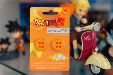 Das Bild zeigt eine Verpackung mit Dragon Ball Z Thumb Grips für den PS5-Controller. Die Grips sind leuchtend gelb und mit einem Design versehen, das vier rote Sterne auf einem gelben Hintergrund zeigt, was sich an den ikonischen Dragonballs orientiert. Im Hintergrund sind verschiedene Figuren und Objekte aus der Dragon Ball Welt zu sehen, darunter eine Figur auf einem motorisierten Roller, was dem Bild einen spielerischen und nerdigen Charakter verleiht. Die Verpackung hebt die Vorteile der Grips hervor, darunter besseren Grip und Präzision beim Spielen.