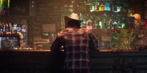 Das Bild zeigt eine Person, die an einer Bar sitzt, und zwar von hinten betrachtet. Die Person trägt ein kariertes Hemd und einen Cowboyhut. Hinter ihnen ist eine bunte Bar mit einer Vielzahl von Flaschen und Getränken, die in unterschiedlichen Farben leuchten. Die Atmosphäre wirkt rustikal und gemütlich, mit vielen dekorativen Elementen und einem leicht vernebelten Raum. Es scheint ein Ort zu sein, der zur Entspannung und zum Genuss von Getränken einlädt.