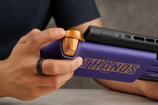 Das Bild zeigt eine Hand, die einen Controller hält, der in auffälligen lila und goldenen Farben gestaltet ist. Der Controller hat das Wort THANOS prominent auf der Seite. Die Hand ist leicht nach oben gedrückt, während das Benutzeroberfläche des Controllers sichtbar ist. Der Hintergrund ist in einem hellen Blau gehalten, was einen frischen und modernen Eindruck vermittelt. Der Fokus liegt auf den ergonomischen Details des Controllers und der Handhaltung, die darauf hindeutet, dass der Controller gerade benutzt wird.