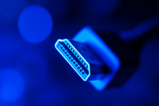 Das Bild zeigt einen fokussierten Ausschnitt eines HDMI-Steckers, beleuchtet von einem blauen Licht, das eine technische und futuristische Atmosphäre schafft. Der Stecker selbst hat eine charakteristische Form mit mehreren Pins, die auf die Verbindungstechnologie hinweisen. Im Hintergrund sind verschwommene, leuchtende Punkte in verschiedenen Farben sichtbar, die dem Bild eine dynamische und lebendige Note verleihen. Diese Darstellung könnte die Wichtigkeit und Funktionalität von HDMI-Kabeln in der Gaming-Welt symbolisieren.