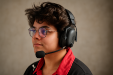 Das Bild zeigt eine Person mit einem Headset auf dem Kopf, das Teil der Razer BlackShark V3 Pro-Serie ist. Die Person hat schwarzes, lockiges Haar und trägt eine rote Jacke mit einem schwarzen Oberteil. Der Hintergrund ist in einem hellen Blau gehalten, was eine lebendige und ansprechende Atmosphäre schafft. Das Headset wirkt modern und professionell, was zum Gaming-Kontext des Bildes passt.