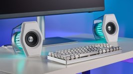 Das Bild zeigt ein modernes Setup mit zwei Lautsprechern, die ein futuristisches Design aufweisen. Die Lautsprecher sind in einem klaren Gehäuse mit einer blauen LED-Beleuchtung, die dynamisch leuchtet. In der Mitte befindet sich eine schlichte, elegante Tastatur mit silbernen Tasten, die gut zu den Lautsprechern passt. Der Hintergrund ist in sanften Blau- und Grautönen gehalten, was dem ganzen Arrangement eine coole Ästhetik verleiht. Dieses Setup scheint besonders für Gamer oder Technikliebhaber geeignet zu sein, die Wert auf sowohl Design als auch Audioqualität legen.