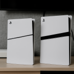 Das Bild zeigt zwei PlayStation 5 Konsolen, die nebeneinander auf einem Tisch stehen. Die Konsolen haben ein modernes, schlankes Design in weiß mit schwarzen Akzenten, und das bekannte PlayStation-Logo ist sichtbar. Im Hintergrund ist ein dunkler Bildschirm zu erkennen, der nicht weiter im Detail erkennbar ist. Der Tisch scheint ordentlich zu sein und es gibt einen weiteren, unscheinbaren Gegenstand im Hintergrund, der sich unterhalb der Konsolen befindet. Die gesamte Komposition vermittelt ein Gefühl von technischer Eleganz und modernen Gaming-Umgebungen.