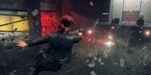 Das Bild zeigt eine actionreiche Szene aus dem Spiel Control. Die Protagonistin bewegt sich dynamisch durch eine Umgebung, in der Materialien und Objekte durch die Luft fliegen. Sie trägt eine dunkle Lederjacke und zielt mit einer Waffe in der Hand, während im Hintergrund rote Lichter und verschwommene Gestalten sichtbar sind. Die Atmosphäre ist spannungsgeladen und vermittelt ein Gefühl von Übernatürlichkeit, das typisch für die Spielwelt ist. Um sie herum sind Partikel und Trümmer zu sehen, die den Eindruck von Bewegung und Chaos verstärken.