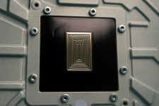 Das Bild zeigt einen Ausschnitt des Innenlebens einer PS5-Konsole, speziell den Bereich rund um die CPU und GPU. In der Mitte ist ein rechteckiges, graviertes Metallstück zu sehen, das offenbar die Liquid-Metal-Wärmeleitpaste beherbergt. Die Struktur weist feine Linien und Rillen auf, die für eine bessere Haftung und eine verbesserte Dichtwirkung sorgen sollen. Um dieses Mittel herum sind mehrere Schrauben sichtbar, die Teile des Gehäuses miteinander verbinden. Der Hintergrund hat eine metallische Anmutung, die die technische Komplexität der Konsole unterstreicht.