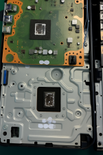 Das Bild zeigt das Innere einer PlayStation 5 Konsole, die geöffnet wurde. Man erkennt die grüne Hauptplatine mit verschiedenen elektronischen Komponenten sowie den Prozessorblock in der Mitte. Rund um diesen Block sind Spuren von Wärmeleitpaste sichtbar, insbesondere im Bereich, wo das liquid metal angewendet wurde. Der Untergrund der Konsole ist ebenfalls sichtbar, mit einer glatten, metallischen Oberfläche. Die gesamte Darstellung vermittelt einen technischen Einblick in die Kühltechnologie der PS5.