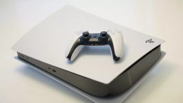 Das Bild zeigt eine PlayStation 5 Slim-Konsole, die in einem eleganten Design in Weiß und Schwarz gehalten ist. Auf der Konsole liegt ein Controller, der ebenfalls in ähnlichen Farben gestaltet ist. Die Konsole hat eine glatte Oberfläche und weist eine markante Form auf, die modern wirkt. Beide Geräte sind auf einem neutralen, einfarbigen Hintergrund platziert, was den Fokus auf die Hardware lenkt. Die gesamte Anordnung vermittelt einen Eindruck von High-Tech und ästhetischer Anziehungskraft im Gaming-Bereich.
