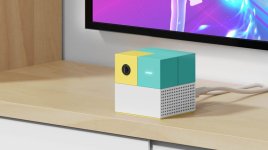 Das Bild zeigt eine kompakte, quadratische Konsole mit einem bunten Design aus hellblauen, gelben und weißen Elementen. Sie steht auf einer Holzoberfläche und ist an ein Fernsehgerät angeschlossen. Der obere Teil der Konsole besteht aus mehreren farbigen Blöcken, während die Unterseite eine strukturierte Oberfläche mit Belüftungslöchern aufweist. Die Konsole wirkt modern und ansprechend, ideal für Familien und Kinder, die Freude an Bewegungsspielen haben.