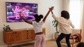 Das Bild zeigt eine Mutter und ihr Kind, die in einem hellen Wohnzimmer aktiv spielen. Sie stehen vor einem Fernseher, auf dem ein Spiel mit dem Titel Barbie angezeigt wird. Beide haben ihre Arme hochgehoben und scheinen die Bewegungen des Spiels nachzuahmen, was auf einen interaktiven Spielstil hindeutet. Der Raum ist freundlich und gut beleuchtet, mit einem modernen Sofa und einem Holzschrank im Hintergrund. Die Atmosphäre wirkt fröhlich und familiär, wobei der Fokus klar auf Bewegung und gemeinsamem Spaß liegt.