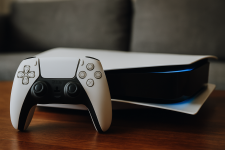 Das Bild zeigt eine PlayStation 5 mit einem weißen Controller, der darauf liegt. Die Konsole hat ein modernes Design mit glatten Linien und einer breiten Form. Der Controller, ebenfalls in weiß, wirkt ansprechend mit seinen charakteristischen Tasten und Joysticks. Der Hintergrund ist einfach gehalten, was den Fokus auf die Konsole und den Controller lenkt. Die gesamte Darstellung vermittelt eine aufregende Atmosphäre für Gamer, besonders im Hinblick auf kommende Spiele wie GTA 6.
