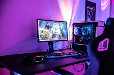 Das Bild zeigt einen modernen Gaming-Setup, bestehend aus einem schlanken Monitor, einer leistungsstarken Gaming-PC-Einheit und einer ansprechenden Tastatur. Die Atmosphäre wird durch sanfte, bunte LED-Lichter in Pink- und Blautönen unterstrichen, die die Wand im Hintergrund erhellen. Auf dem Schreibtisch liegt ein headset mit bunten Elementen, während der Stuhl ergonomisch gestaltet ist und Komfort beim Spielen bietet. Alles wirkt stilvoll und einladend für Gamer und Streamer.