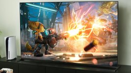 Das Bild zeigt eine moderne Spielkonsole, die PS5, neben einem Fernseher, der ein lebhaftes Bild aus dem Spiel Ratchet & Clank: Rift Apart zeigt. Auf dem Bildschirm sieht man den Charakter Ratchet, der eine futuristische Waffe abfeuert, während Energiestrahlen durch die Luft schießen. Die Szene ist farbenfroh und dynamisch, was die aufregende Action des Spiels widerspiegelt. Die Umgebung wirkt stilvoll und modern, passend zur Optik der Konsole. Ein Controller liegt griffbereit in der Nähe, bereit für das nächste Gaming-Abenteuer.