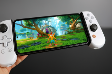Das Bild zeigt eine Hand, die ein Smartphone mit einem angeschlossenen Controller hält. Auf dem Bildschirm des Smartphones ist ein farbenfrohes Videospiel zu sehen, das eine lebendige Landschaft mit Bäumen und einer Spielfigur zeigt. Die Controller umfassen die Seiten des Smartphones und verfügen über Tasten und Analogsticks, die für das Spielen von Videospielen genutzt werden. Der Hintergrund ist unscharf, wodurch der Fokus auf dem Controller und dem Bildschirm liegt. Dieses Setup ermöglicht es, Spiele bequem von unterwegs zu spielen.