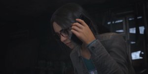 Das Bild zeigt eine junge Frau, die in einem leicht düsteren Raum steht und ein Smartphone hält. Sie hat mittellange, glatte Haare und trägt eine Brille. Ihr Gesichtsausdruck wirkt konzentriert, während sie in ein Telefon spricht. Der Hintergrund ist unscharf, aber man erkennt einige Möbelstücke und Fenster, die eine unheimliche Atmosphäre erzeugen. Die Beleuchtung ist dämmrig und trägt zur Spannung der Szene bei.