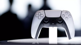 Das Bild zeigt einen modernen Spiel-Controller, der für die PlayStation 5 entwickelt wurde. Er steht auf einer weißen Oberfläche und hebt sich durch sein futuristisches Design und die Kombination aus Schwarz und Weiß ab. Im Hintergrund sind unscharfe Silhouetten sichtbar, was dem Bild eine dynamische und technische Atmosphäre verleiht. Der Controller ist so positioniert, dass er im Vordergrund auffällt und die Aufmerksamkeit auf seine Form sowie die Bedienelemente lenkt.