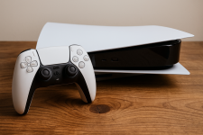 Das Bild zeigt eine PlayStation 5 Konsole, die auf einem Holztisch platziert ist. Neben der Konsole liegt ein DualSense Controller. Die PS5 hat ein modernes, elegantes Design in weiß und schwarz, mit abgerundeten Kanten. Der Tisch hat eine natürliche Holzmaserung, die einen warmen Kontrast zur technologischen Ästhetik der Spielkonsole bildet. Das Licht fällt sanft auf die Oberfläche und unterstreicht die Details des Designs.