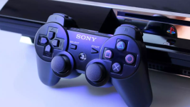 Das Bild zeigt einen PlayStation 3 Controller in naher Ansicht, der auf einem Tisch liegt, vor einem Teil der Konsole. Der Controller ist schwarz mit typischen PlayStation-Tasten, darunter die bekannten Symbole für verschiedene Funktionen. Die Beleuchtung sorgt für einen dezenten Glanz, der dem gesamten Bild eine moderne und technologische Atmosphäre verleiht. Die Kombination aus Controller und Konsole weckt nostalgische Erinnerungen an die Gaming-Zeit vergangener Tage, während gleichzeitig der Bezug zur aktuellen Diskussion um Retro-Games hergestellt wird.