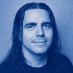 Das Bild zeigt einen Mann mit langen, dunklen Haaren, der direkt in die Kamera schaut. Der Hintergrund ist in einem hellen Farbton gehalten, möglicherweise um einen modernen und klaren Eindruck zu vermitteln. Der Gesichtsausdruck des Mannes wirkt freundlich und offen, was zusammen mit dem simplen Hintergrund eine zugängliche und professionelle Atmosphäre schafft.